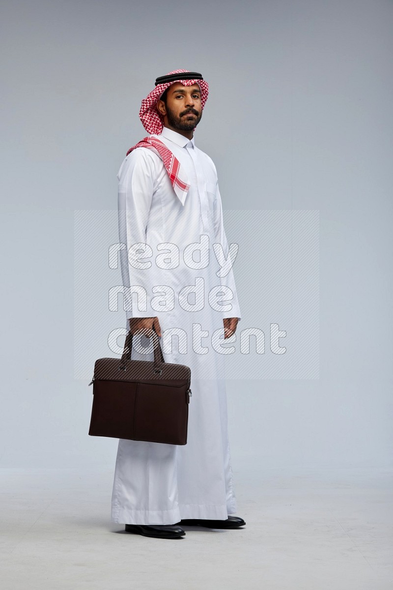 Saudi man Wearing Thob and shomag standing holding bag on Gray background