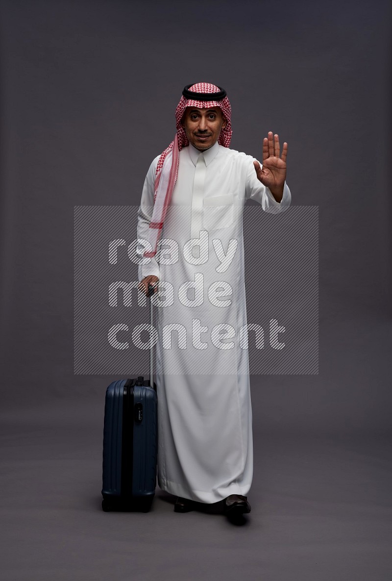 Saudi man wearing thob and shomag standing holding bag on gray background