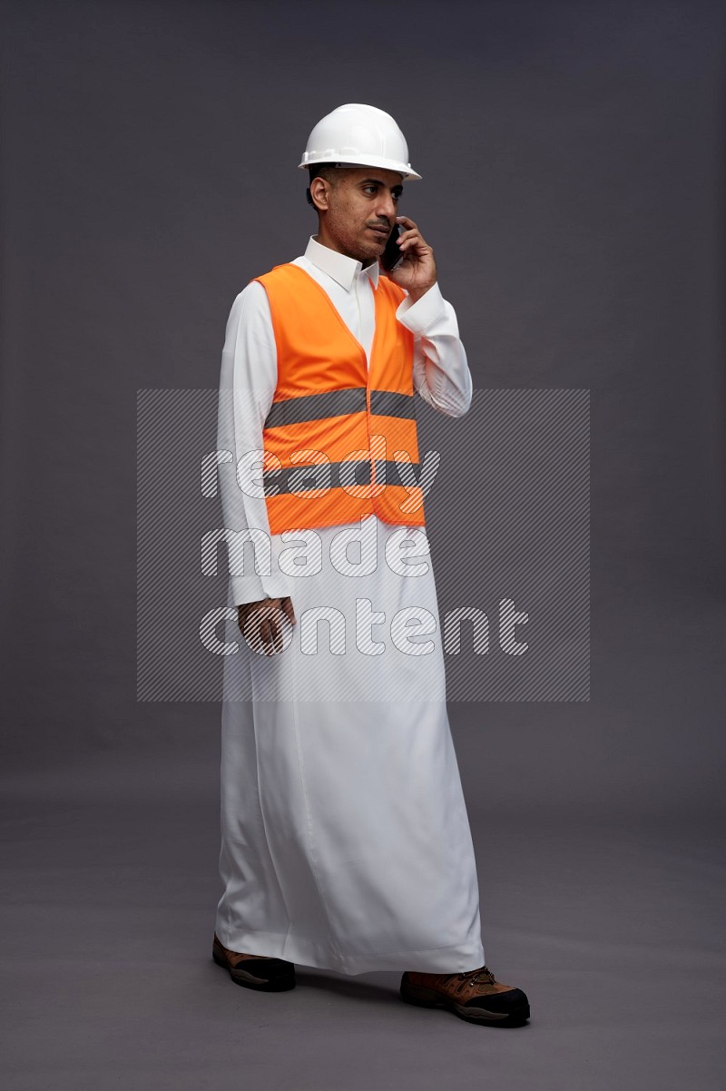 Saudi man wearing thob with engineer vest standing talking on phone on gray background