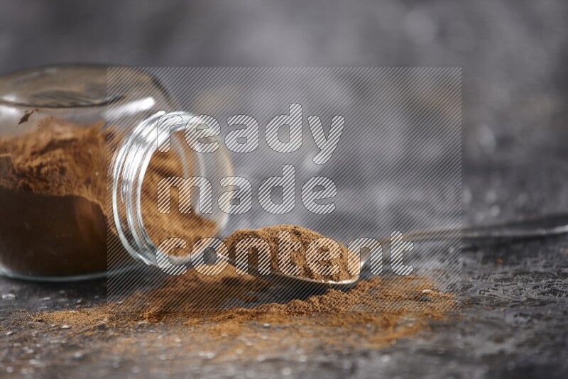 Herbal glass jar full of cinnamon powder flipped and a metal spoon on textured black background