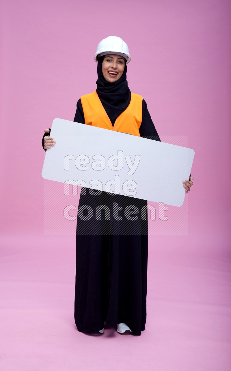 Saudi woman wearing Abaya with engineer vest and helmet standing holding board on pink background