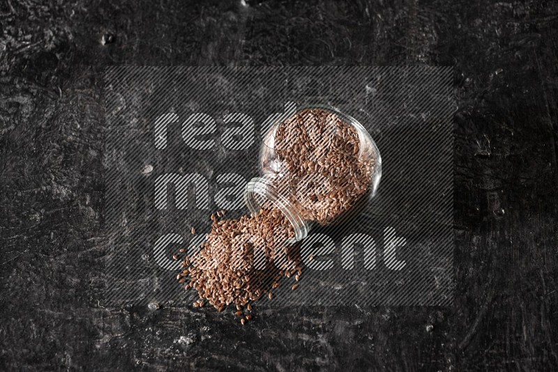 A glass spice jar full of flaxseeds flipped and seeds spread out on a textured black flooring