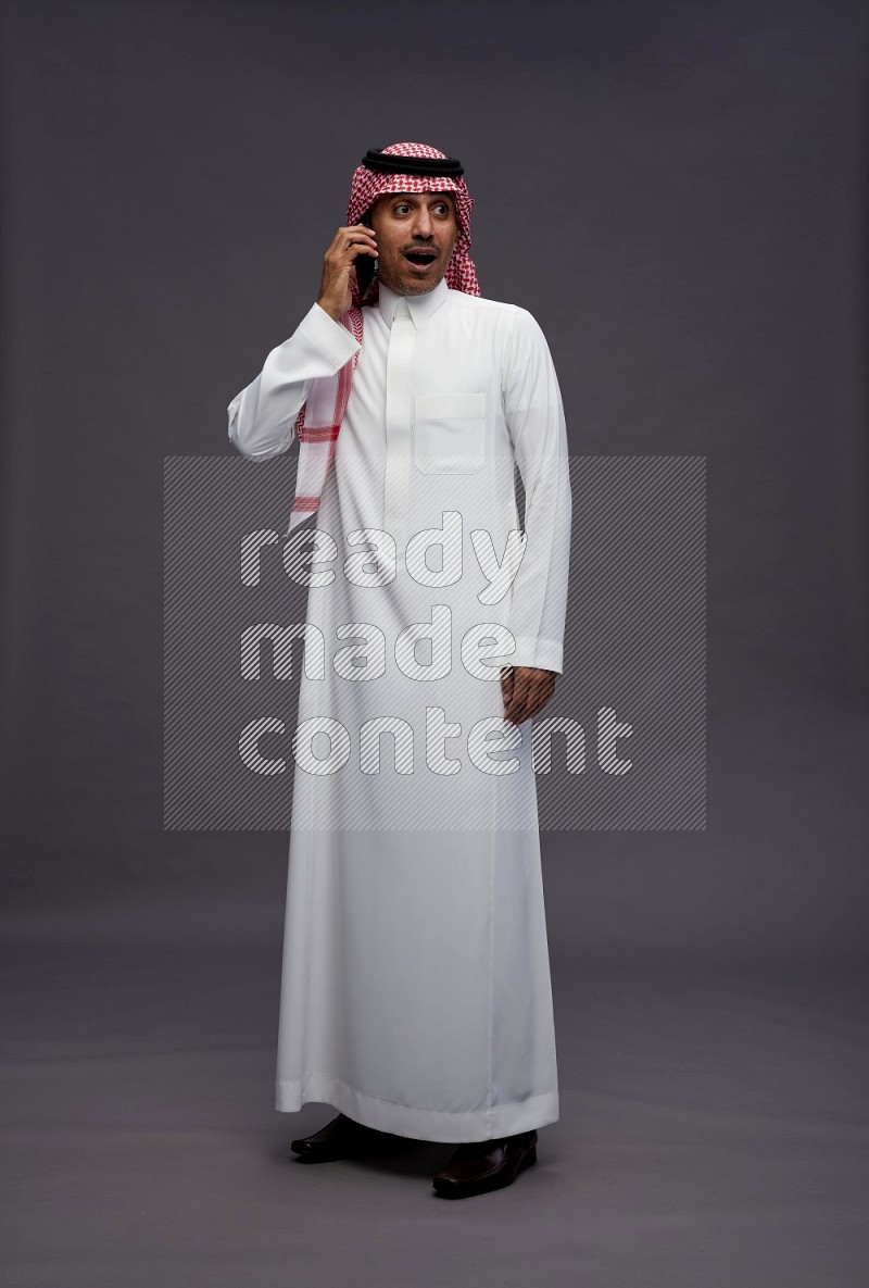 Saudi man wearing thob and shomag standing talking on phone on gray background