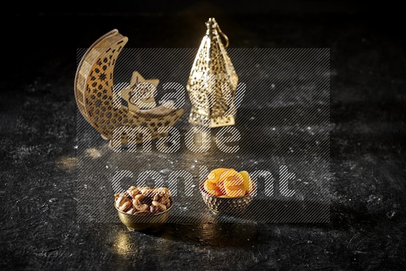Nuts in a metal bowl with dried apricots beside golden lanterns in a dark setup