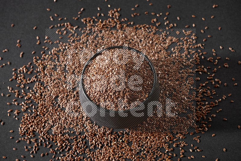 A black pottery bowl full of flaxseeds surrounded by the seeds on a black flooring