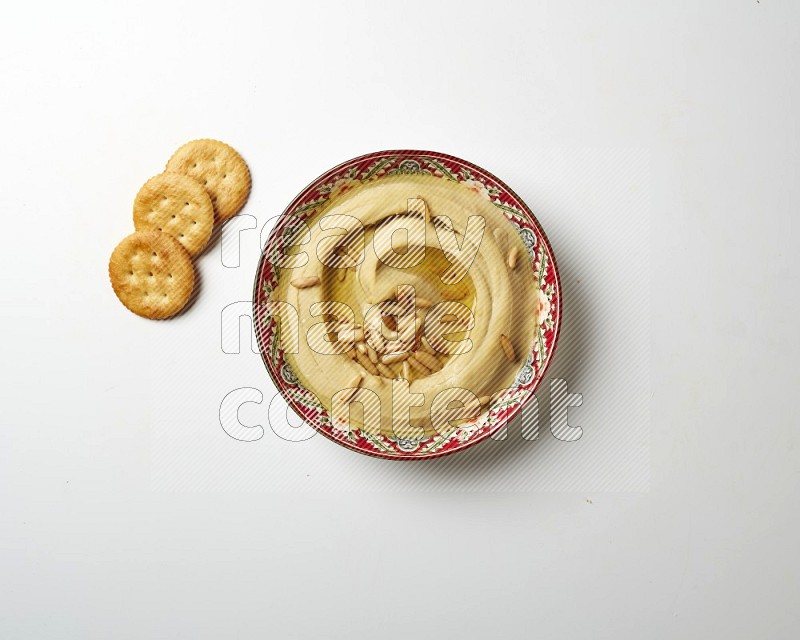 Hummus in a red plate with patterns garnished with pine nuts on a white background