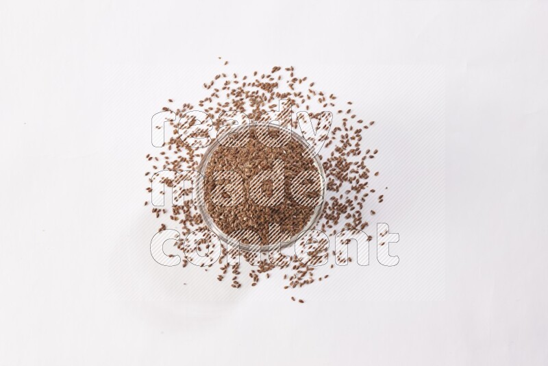 A glass bowl full of flax seeds surrounded by flax seeds on a white flooring