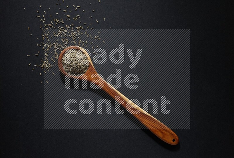 A wooden ladle full of cumin seeds on black flooring