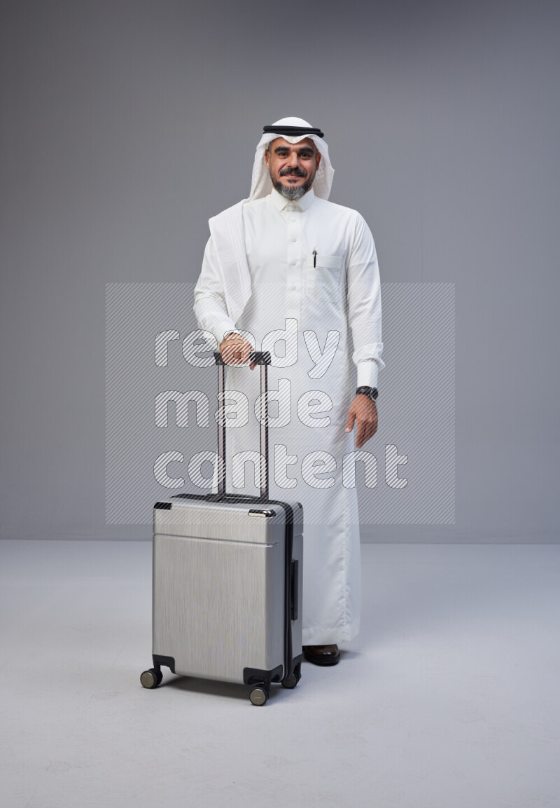 Saudi man wearing Thob and white Shomag standing holding Travel bag on Gray background