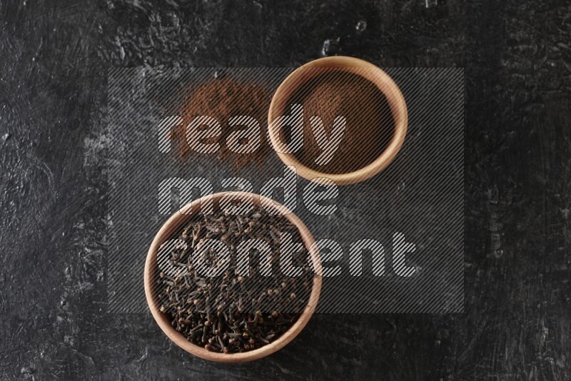 2 wooden bowls full of cloves powder and whole cloves on a textured black flooring