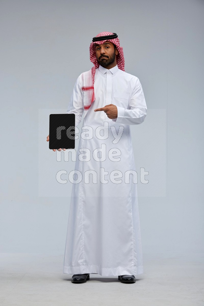 Saudi man Wearing Thob and shomag standing showing tablet to camera on Gray background