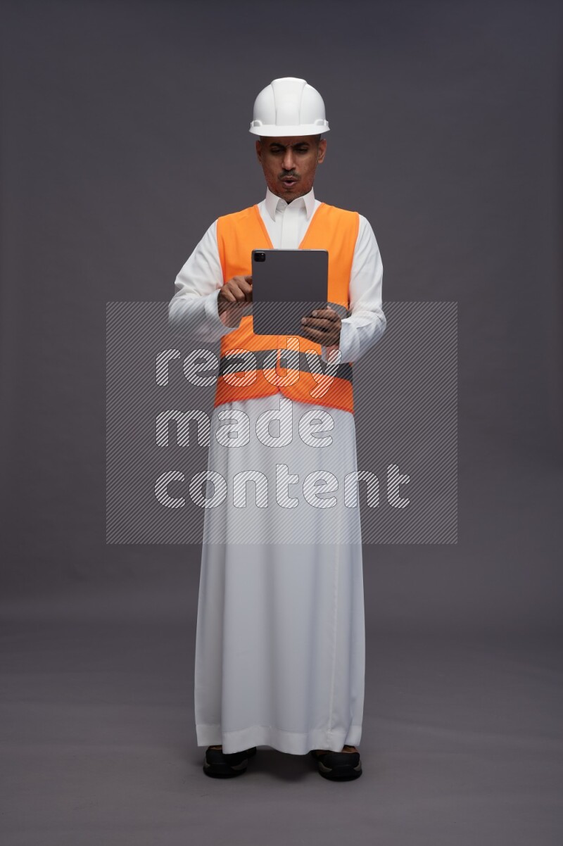 Saudi man wearing thob with engineer vest standing working on tablet on gray background