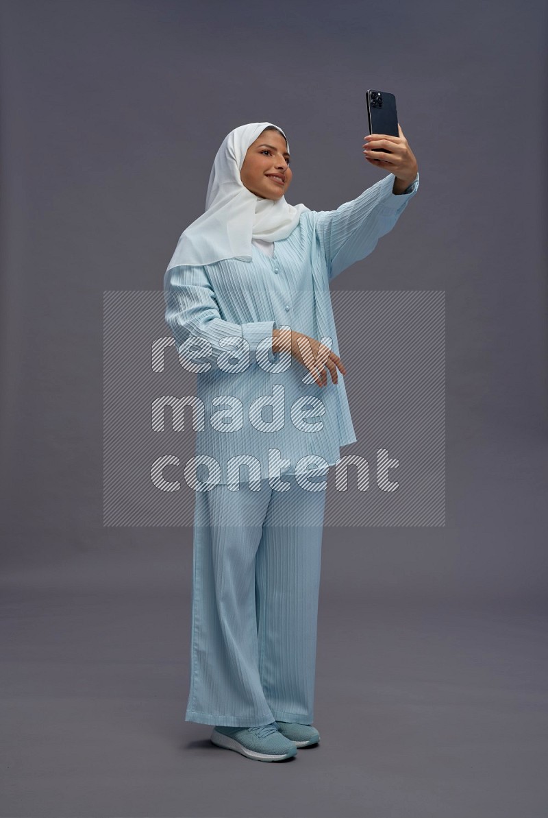 Saudi woman wearing hijab clothes standing taking selfie on gray background