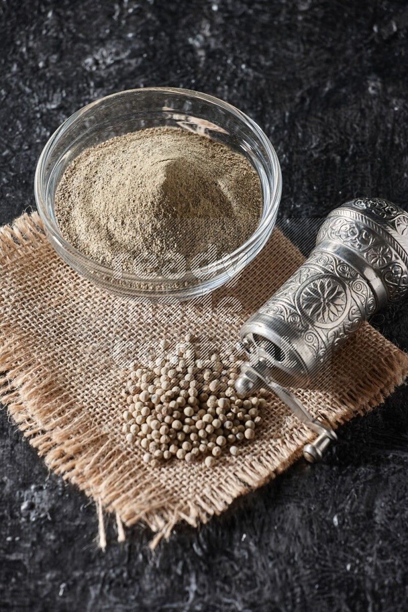 A glass bowl full of white pepper powder with white pepper beads on a burlap piece of fabric and a metal grinder on textured black flooring