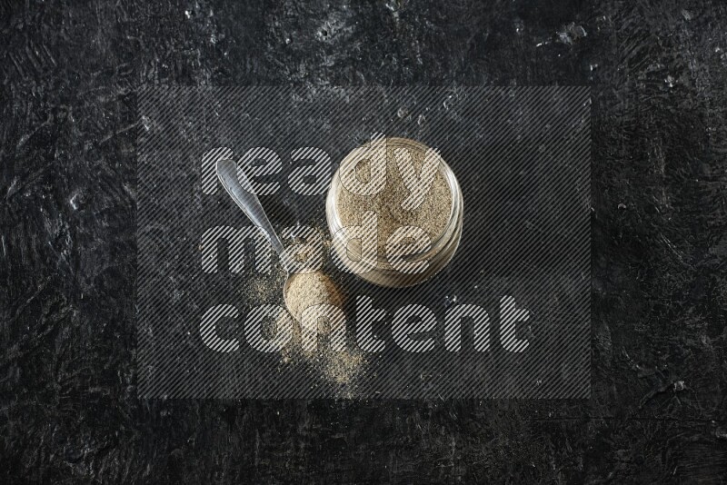 A glass jar and metal spoon full of cardamom powder on textured black flooring