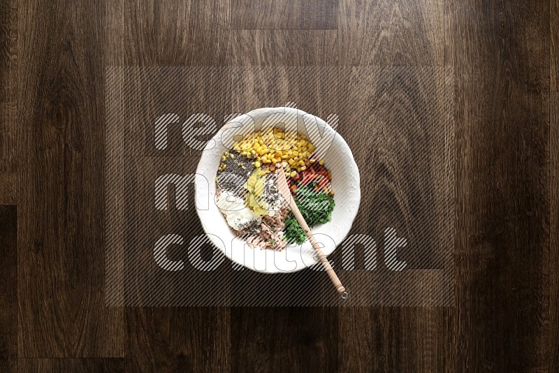 A white bowl full of tuna, colored bell pepper, sour cream, corn, parsley, black pepper powder and sauce, with wooden spoon on wooden background