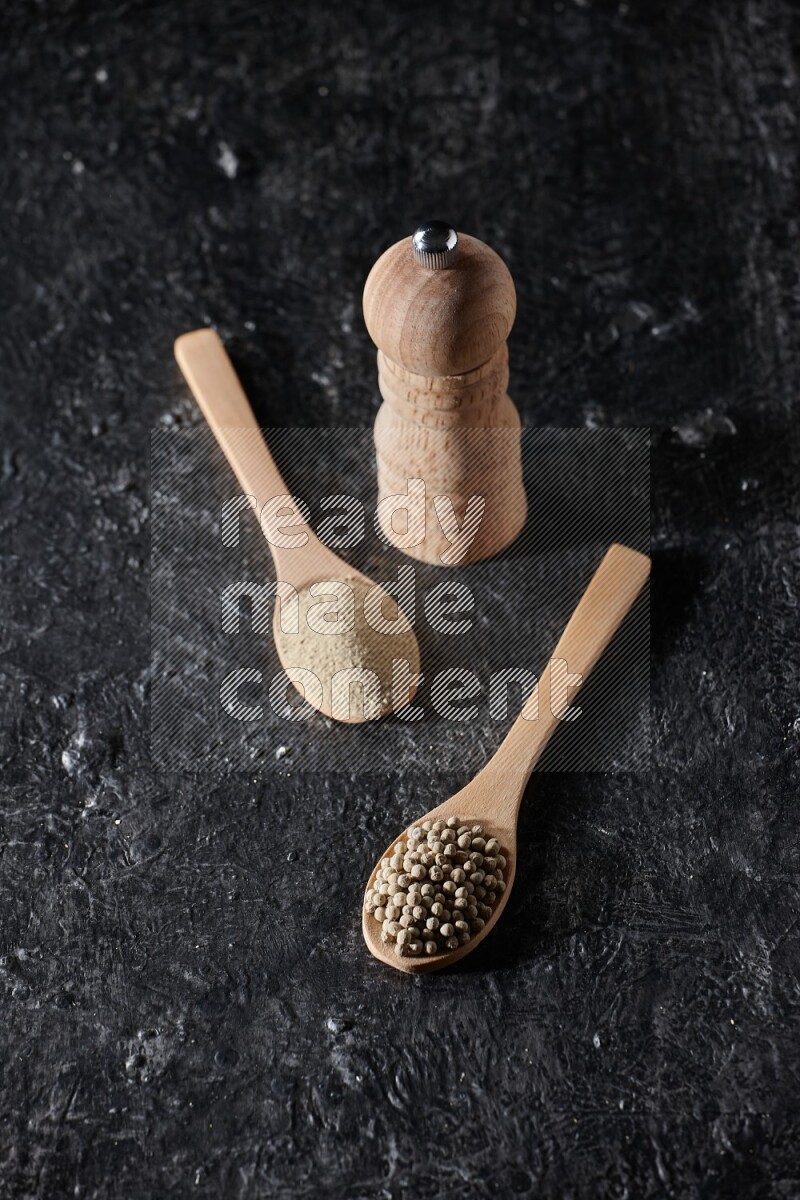 2 wooden spoons one full of white pepper powder and the other with pepper beads and a wooden pepper mill on textured black flooring