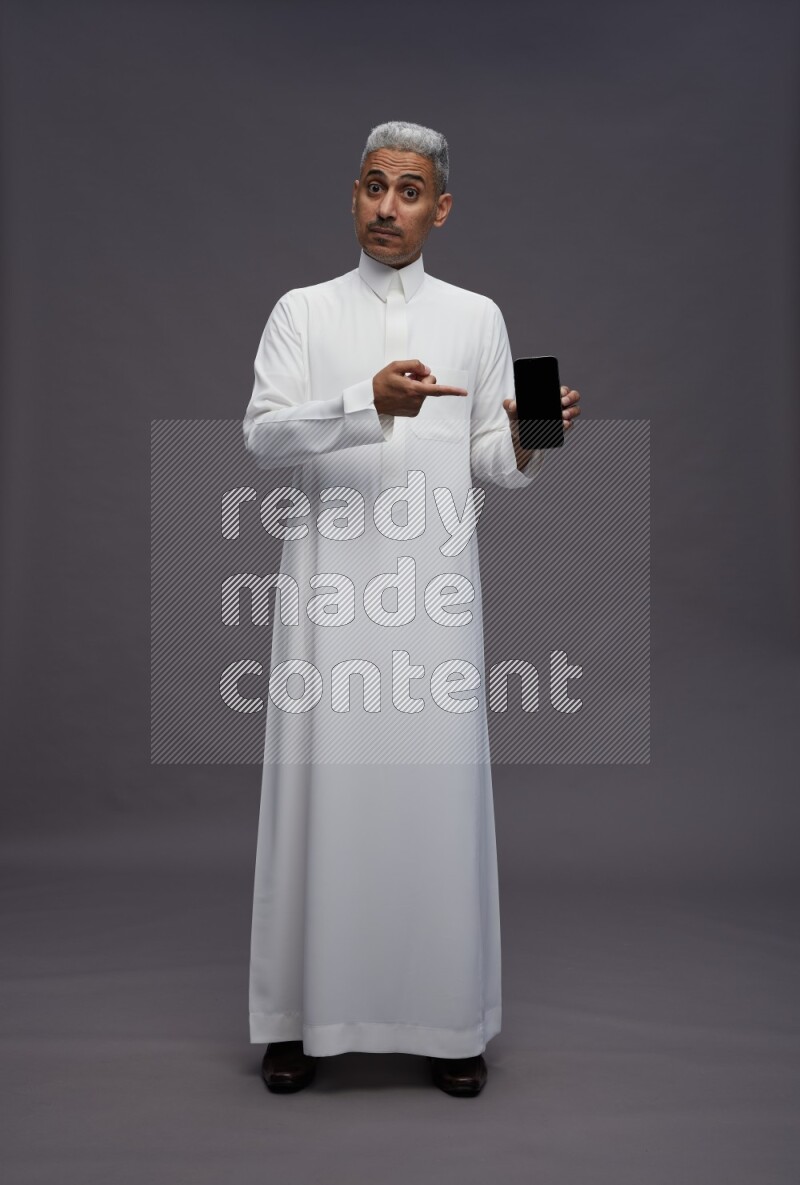 Saudi man wearing thob standing showing phone to camera on gray background
