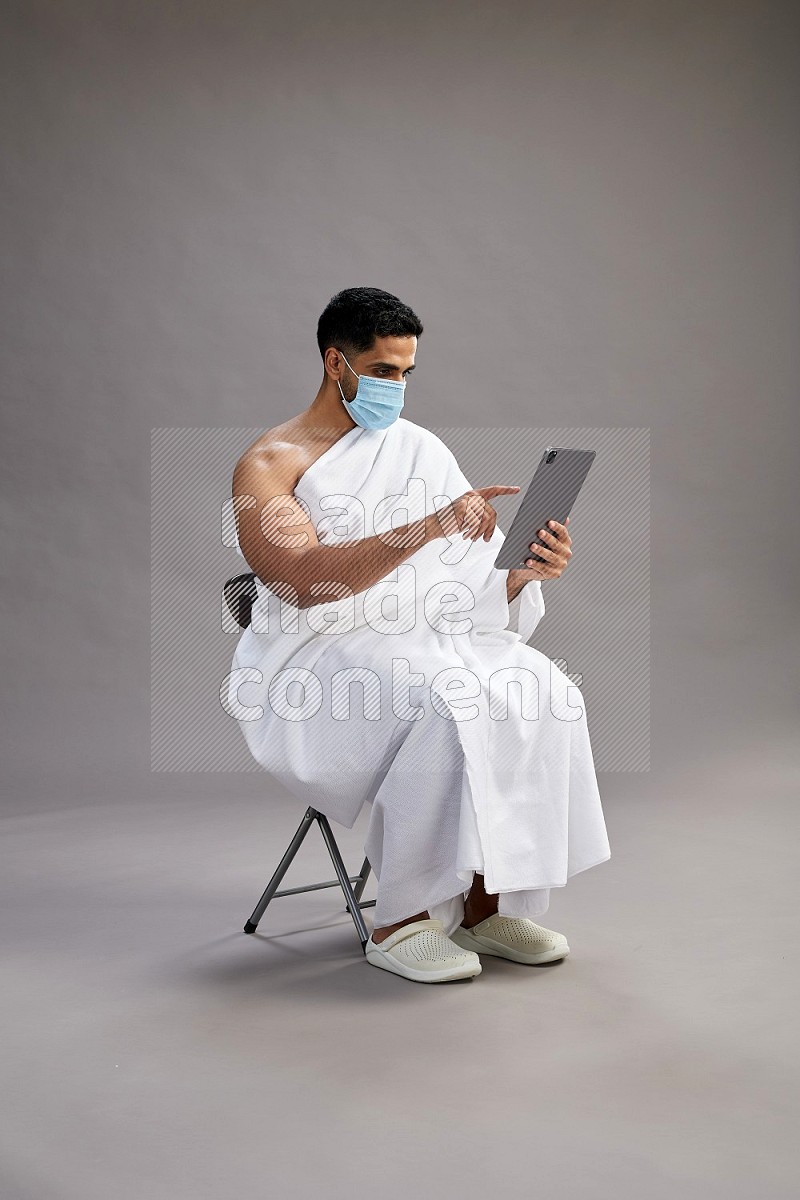 A man wearing Ehram with face mask sitting on chair holding a tablet on gray background