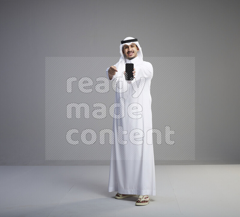 A Saudi man standing wearing thob and white shomag showing phone to camera on gray background
