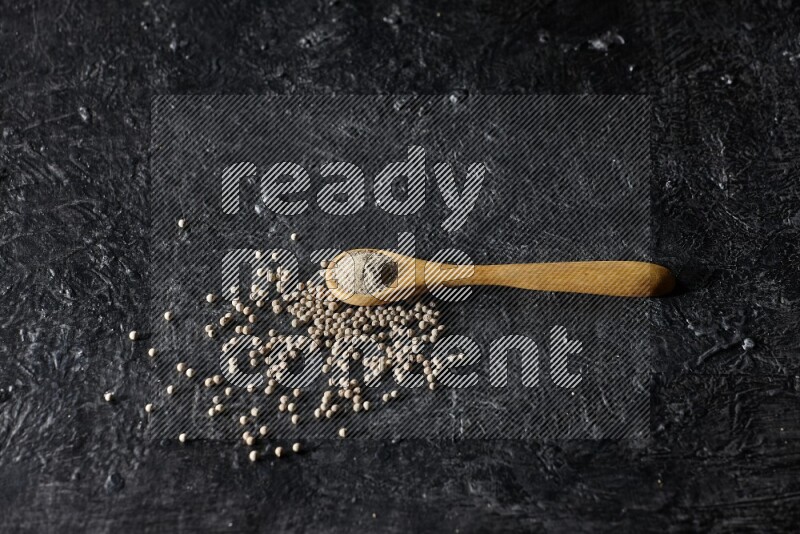 A wooden spoon full of white pepper powder with white pepper beads on textured black flooring