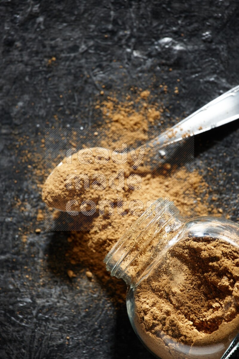 A flipped glass spice jar and a metal spoon full of allspice powder and powder spilled out of it on a textured black flooring