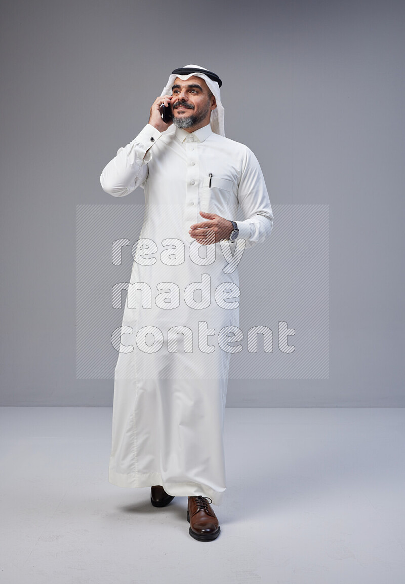 Saudi man Wearing Thob and white Shomag standing talking on phone on Gray background