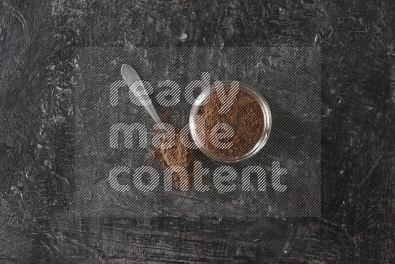 A glass jar full of cloves powder with a metal spoon on a textured black flooring
