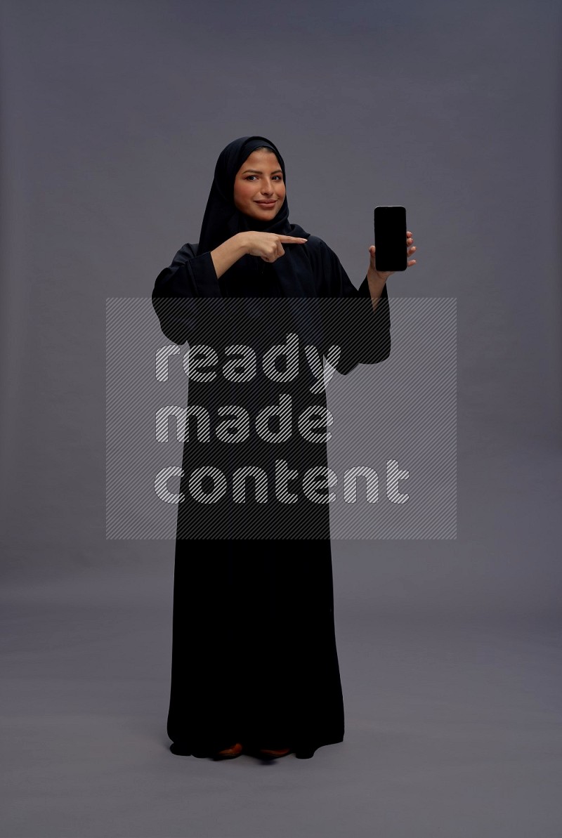 Saudi woman wearing Abaya standing showing phone to camera on gray background