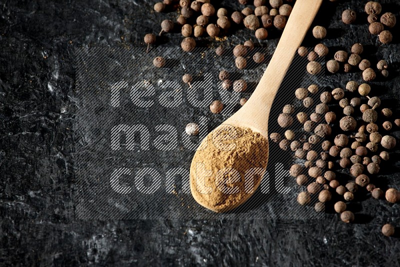 Wooden spoons full of all spice powder and allspice whole balls beside it on a textured black flooring