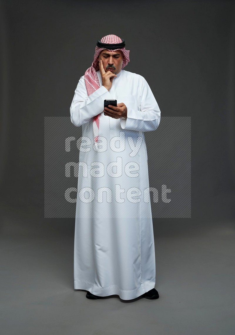 Saudi man with shomag Standing texting on phone on gray background