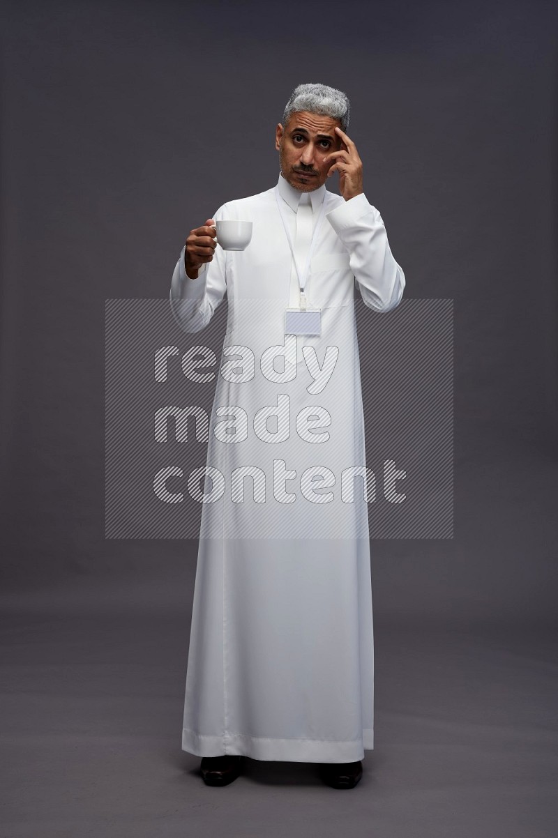 Saudi man wearing thob with neck strap employee badge standing holding mug on gray background