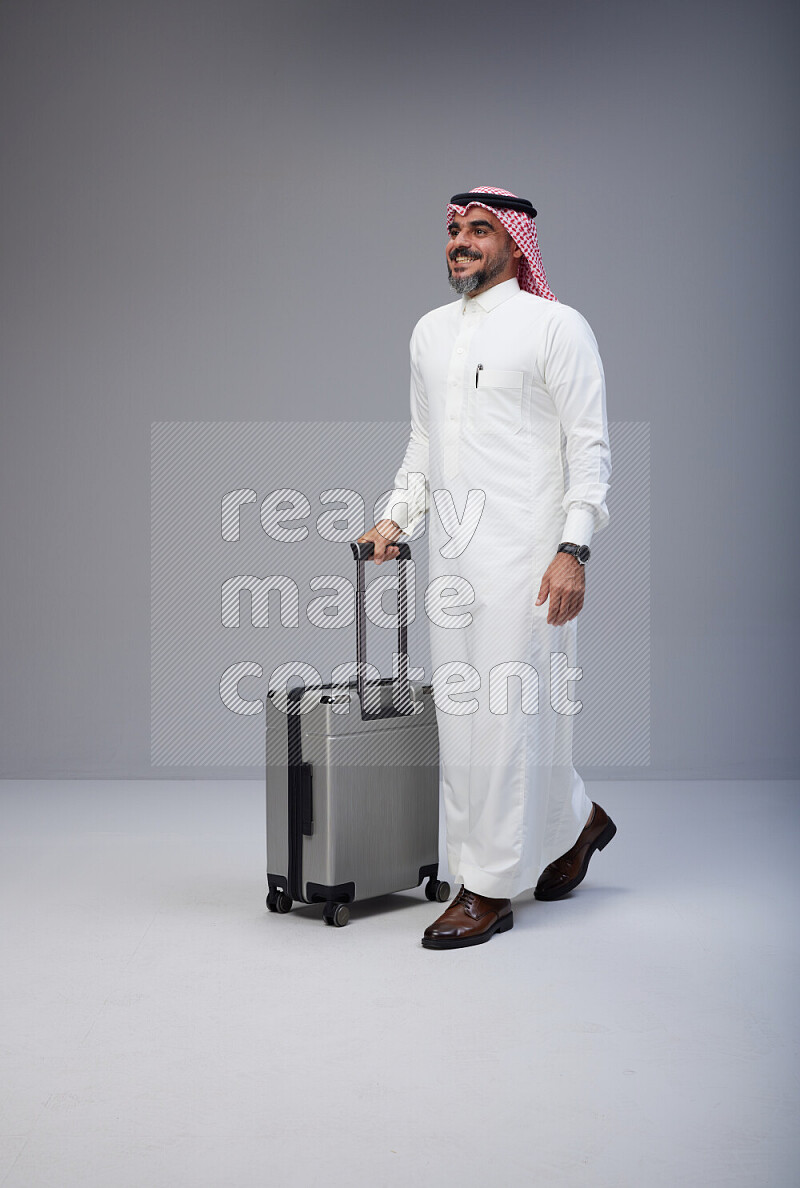 Saudi man wearing Thob and red Shomag standing holding Travel bag on Gray background