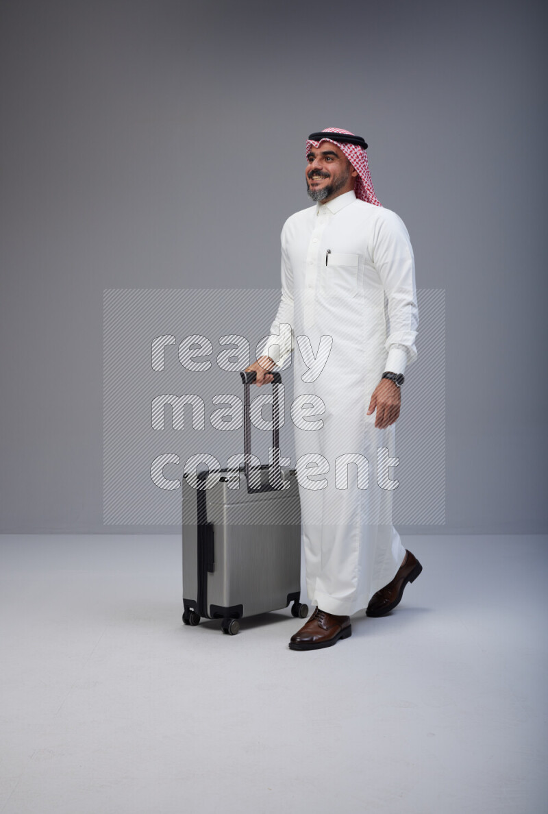 Saudi man wearing Thob and red Shomag standing holding Travel bag on Gray background