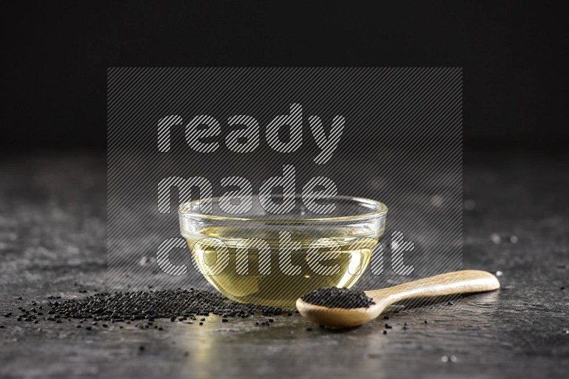 A glass bowl full of black seeds oil and wooden spoon full of black seeds with seeds spreaded on a textured black flooring