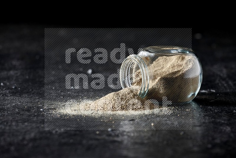 A flipped herbal glass jar full of white pepper powder with spilled powder on textured black flooring