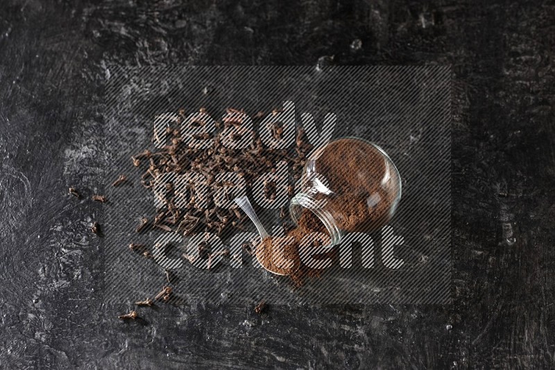 A flipped glass spice jar and a metal spoon full of cloves powder with cloves spread on textured black flooring