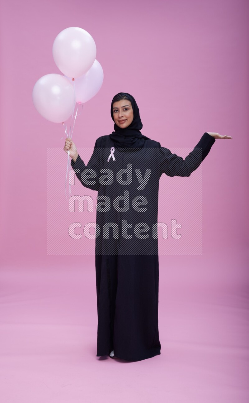 Saudi woman wearing pink ribbon on Abaya standing holding balloons on plain pink background