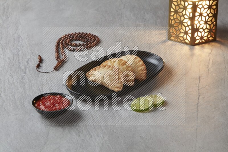 Three fried sambosas in an oval shaped black plate and a red sauce in a black round ramekin with a brown misbaha and a golden lantern on the side on a gray background
