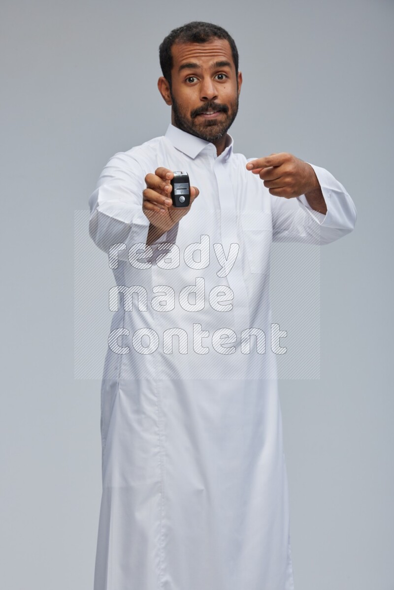 Saudi man wearing thob and shomag standing holding car key on gray background