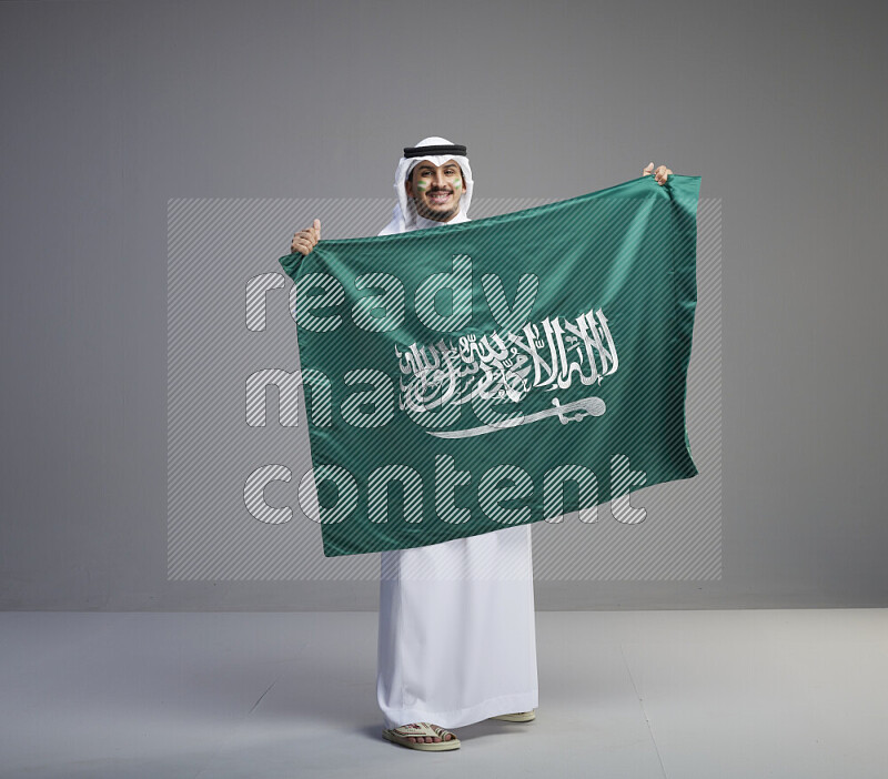 A Saudi man standing wearing thob and white shomag with face painting holding big Saudi flag on gray background