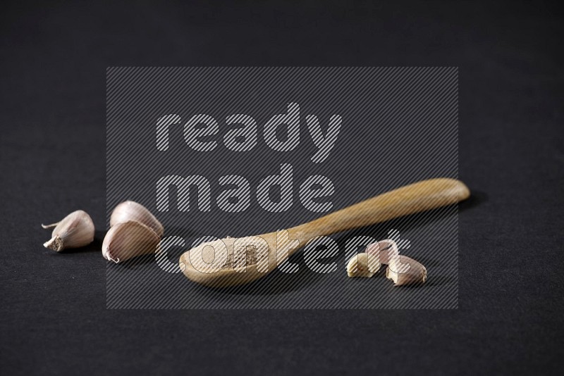 A wooden spoon full of garlic powder with cloves beside it on a black flooring