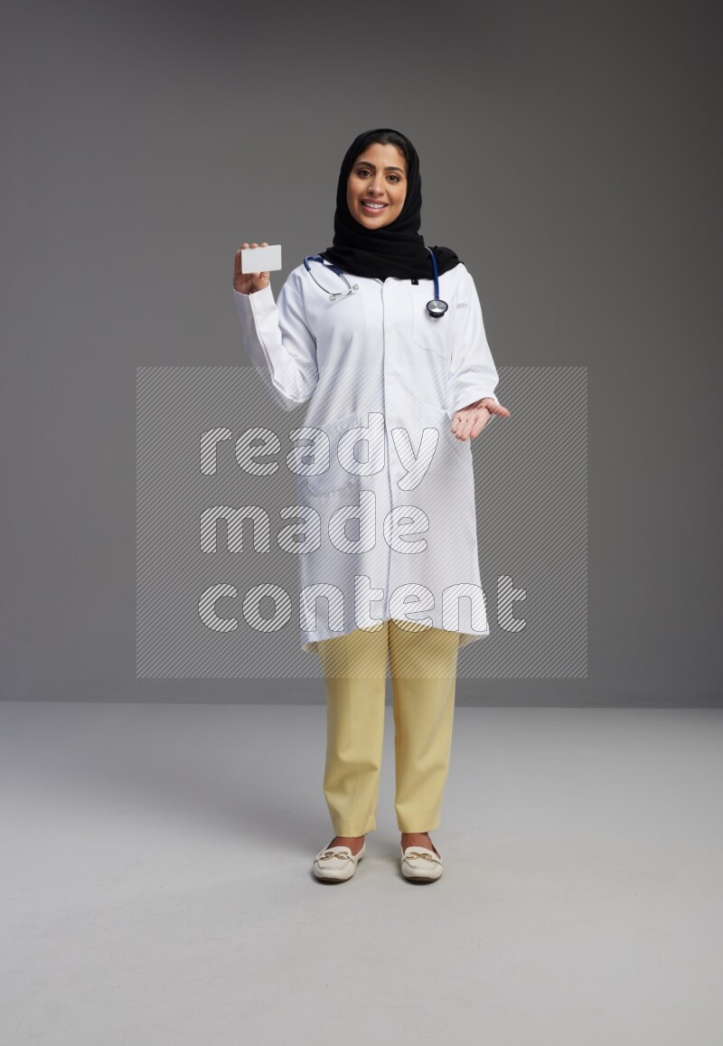 Saudi woman wearing lab coat with stethoscope standing holding ATM card on Gray background