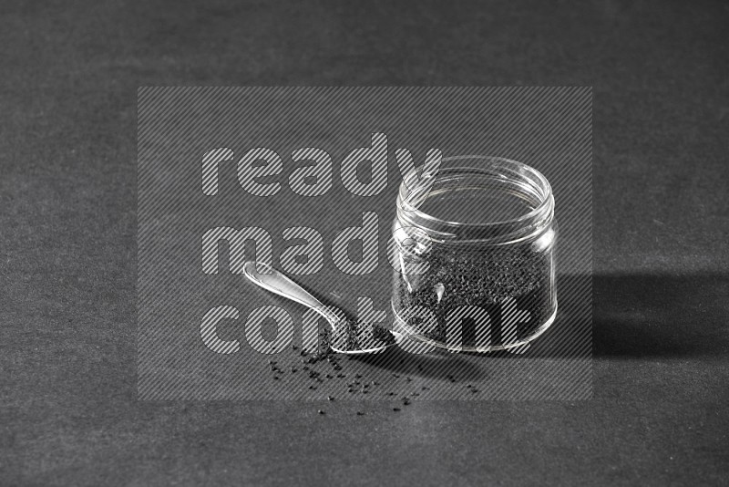 A glass jar full of black seeds with a metal spoon full of the seeds on a black flooring