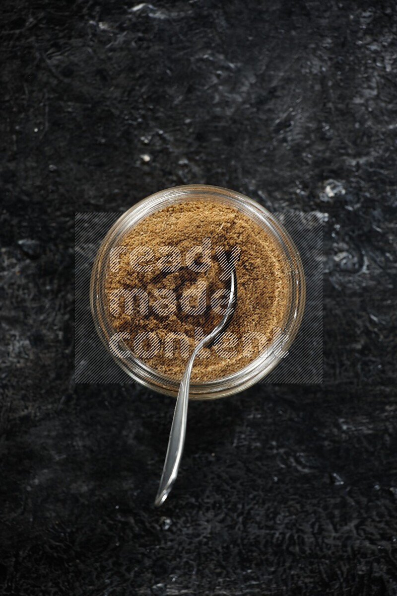 وعاء زجاجي للتوابل وملعقة معدنية ممتلئين ببودرة الكمون على خلفية سوداء