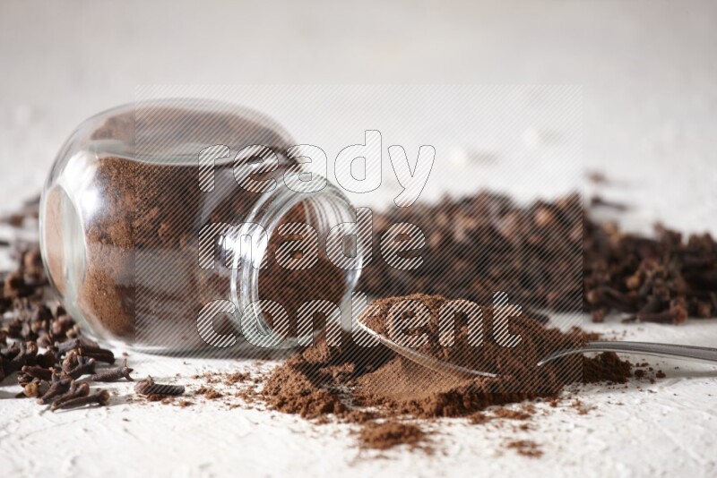 A flipped glass spice jar and a metal spoon full of cloves powder and powder came out of the jar with cloves spread on textured white flooring