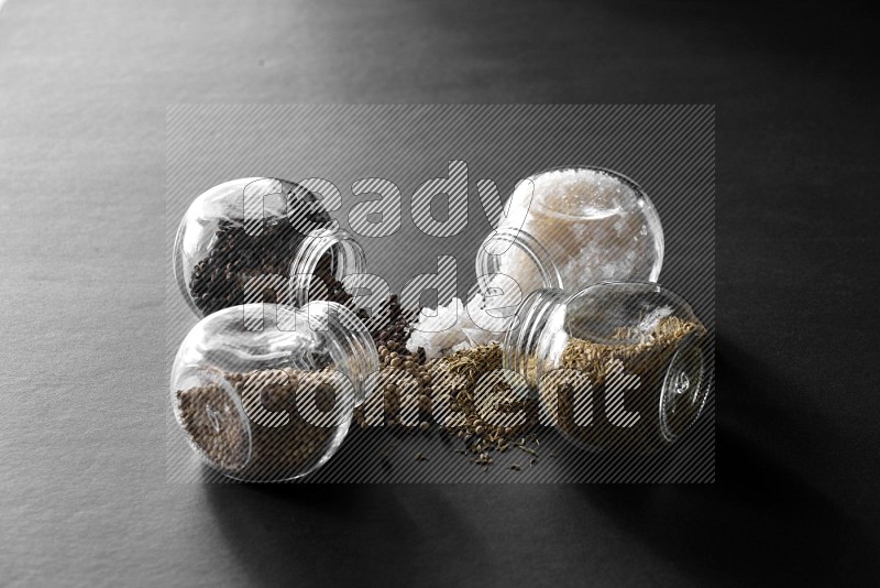 4 glass spice jars full of salt, black peppers, white peppers and cumin on black flooring