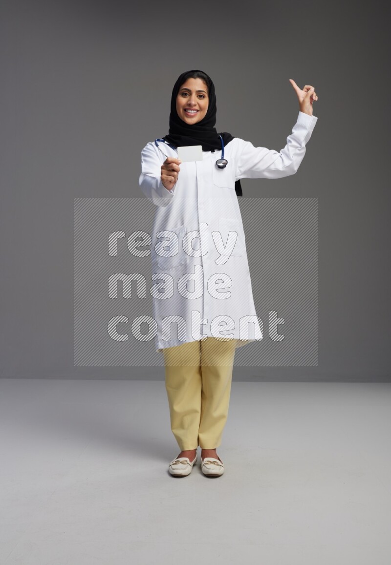 Saudi woman wearing lab coat with stethoscope standing holding ATM card on Gray background