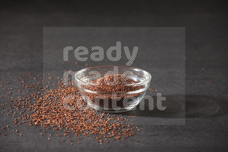 A glass bowl full of garden cress seeds surrounded by seeds on a black flooring