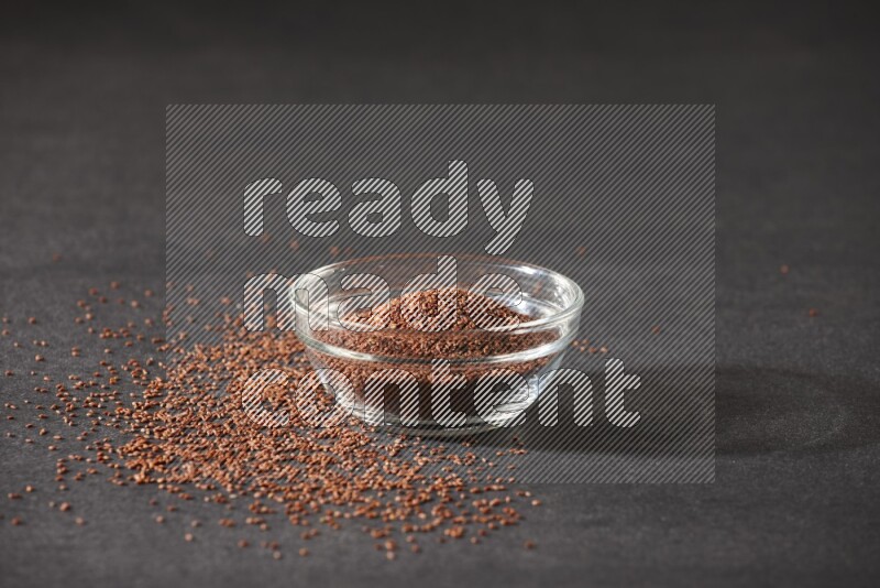 A glass bowl full of garden cress seeds surrounded by seeds on a black flooring